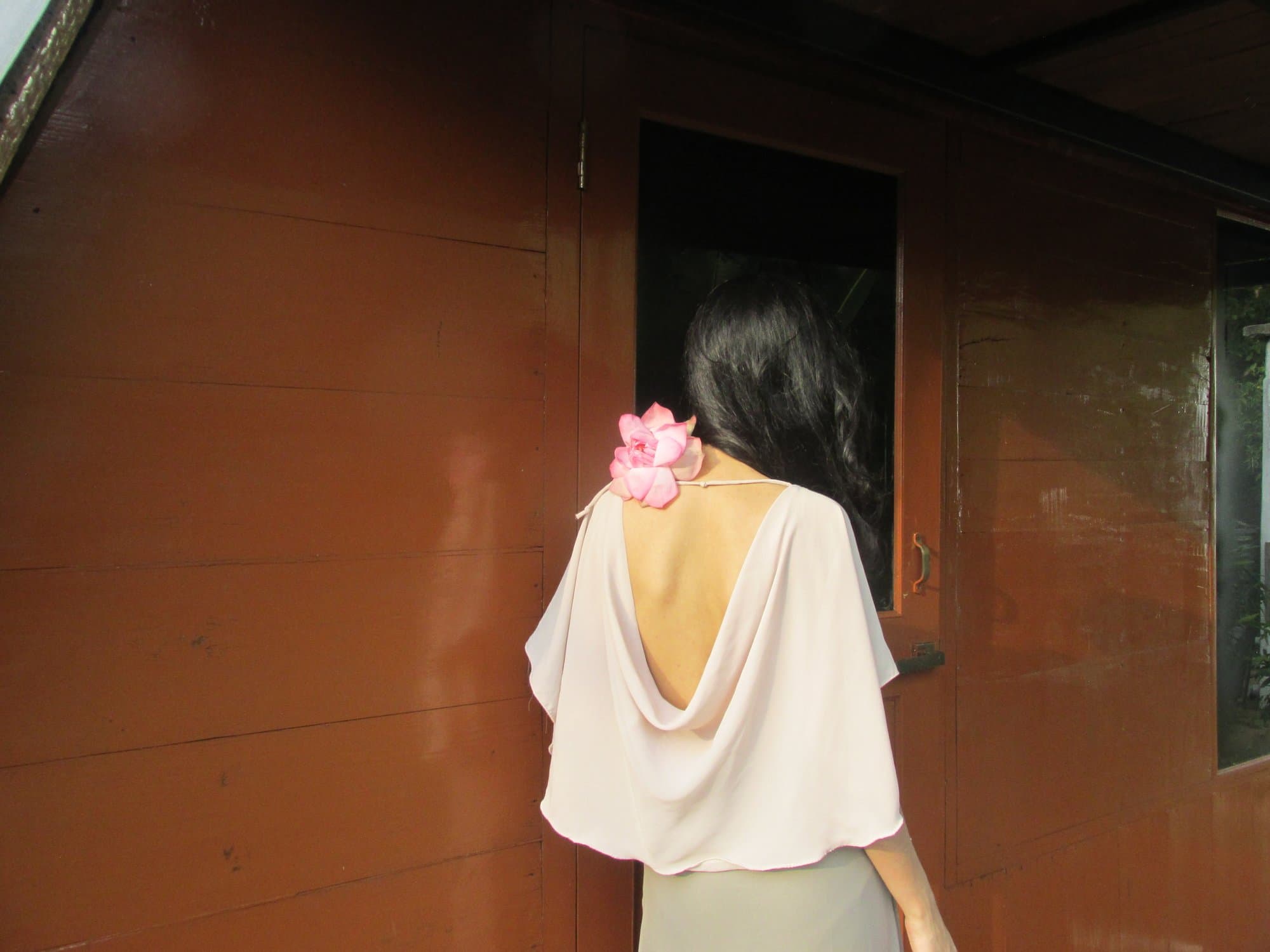 A woman seen from behind in a blush cape, a single pink lotus resting at her bared shoulder.